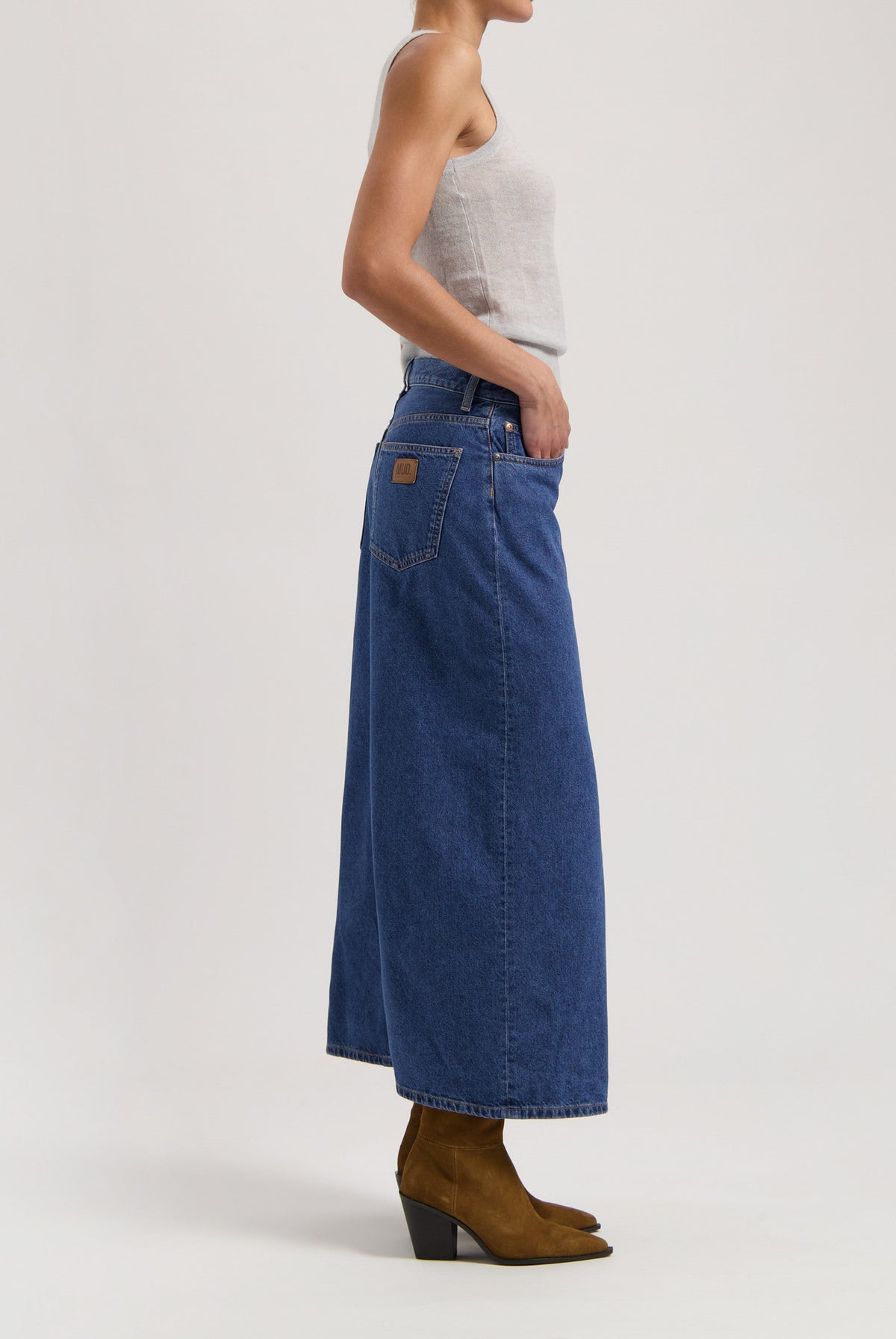 Woman wearing a sleeveless gray top and high-waisted blue denim maxi skirt paired with brown leather ankle boots.